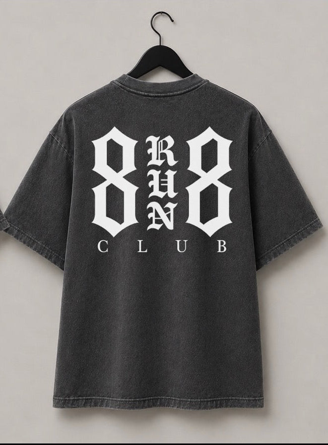Two views of a dark gray washed t-shirt hanging on black hangers against a light gray wall. The front view shows a small white '888 CLUB' logo on the left chest, and the back view shows a large white '888 RUN CLUB' graphic.