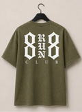 An olive green short-sleeve t-shirt shown from both front and back, hanging on black hangers against a light beige wall. The front features a small white graphic on the left chest, and the back features a large white graphic reading '8 RUN 8 CLUB'.
